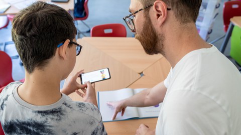 Das Foto zeigt einen Jungen und einen Mann von hinten. Der Mann erklärt dem Jungen etwas und zeigt dabei in ein Buch. Der Junge hält ein Smartphone in der Hand.
