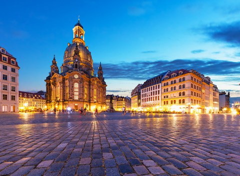 Foto des Dresdner Neumarktes mit der Frauenkirche. Es ist Abend, einige Menschen sind auf dem Neumarkt zu Fuß unterwegs.