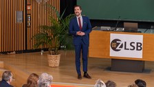 Moderator steht auf der Bühne und spricht zum Publikum.