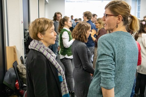 Zwei Frauen, die sich unterhalten. Im Hintergrund herrscht buntes Treiben.
