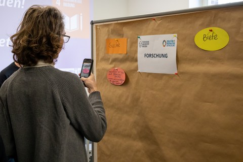 Ein junger Mann fotografiert etwas von einer Pinnwand mit dem Smartphone ab.