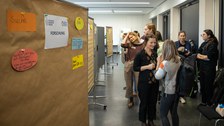 Das Foto zeigt eine Pinnwand mit Personen im Hintergrund, die sich unterhalten.