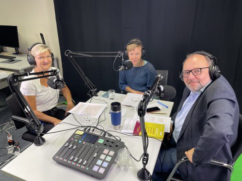 3 Personen sitzen an einem Tisch während einer Podcastaufnahme
