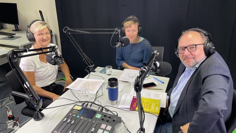 3 Personen sitzen an einem Tisch während einer Podcastaufnahme