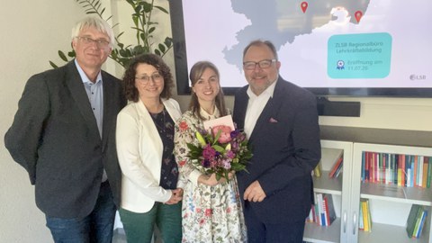 2 Frauen und 2 Männer stehen vor einem großen Bildschirm mit Blumenstrauß