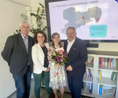 2 Frauen und 2 Männer stehen vor einem großen Bildschirm mit Blumenstrauß