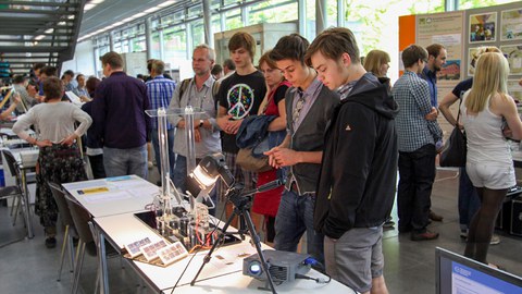 Auf dem Bild sind im Vordergrund zwei Jungs zu sehen, die sich einen Versuchsaufbau ansehen. Im Hintergrund sind einige mehr Menschen zu sehen, die sich den Versuchsaufbau ebenfalls angucken.