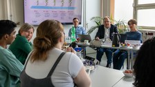 Sechs Personen sitzen sich in jeweils aufgeteilt gegenüber und unterhalten sich, während eine Präsentation im Hintergrund zu sehen ist. 