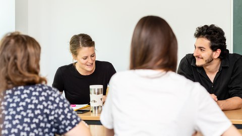 Foto von vier Personen, die sich an zwei Tischen gegenübersitzen und sich unterhalten.