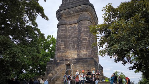 Das Foto zeigt den Bismarckturm in Dresden, vor dem Mitwirkende an der Gedenklesung sitzen und stehen.