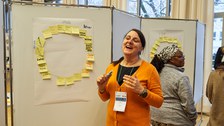 Eine Frau erklärt etwas an einem Whiteboard, an welchem ein Kreis aus Post-its hängt.
