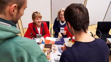 Zwei Frauen sitzen an einem Tisch mit Süßigkeiten und Infomaterial und sprechen mit Studierenden.