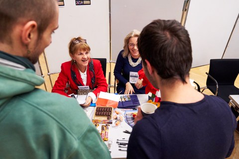 Zwei Frauen sitzen an einem Tisch mit Süßigkeiten und Infomaterial und sprechen mit Studierenden.