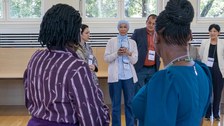 Frau im Hintergrund spricht in ein Mikro. Im Vordergrund sind zwei Frauen zu sehen, die Richtung Hintergrund gucken.