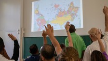 Gruppe von Personen, die im Vordergrund ihre Hände heben. Im Hintergrund ist eine Weltkarte an die Wand projiziert. 