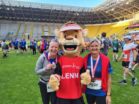 zwei Frauen im Sportoutfit stehen links und rechts neben einem Maskottchen