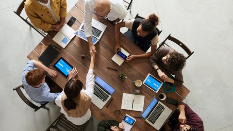 Foto einer Gruppe von 8 Personen in Vogelperspektive. Die Personen sitzen um einen Holztisch und arbeiten an mobilen Endgeräten, Laptops oder mit Notizbüchern. Eine Frau und ein Mann reichen sich über den Tisch hinweg die Hand.