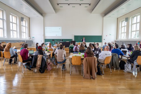 Eine Menschengruppe sitzt in einem Hörsaal und hört einer Präsentation zu.