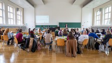 Eine Menschengruppe sitzt in einem Hörsaal und hört einer Präsentation zu.