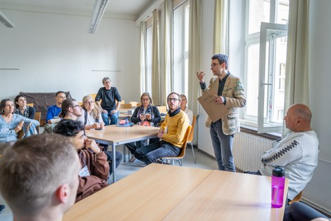 Ein Mann erklärt an Tischen sitzenden Personen etwas.