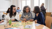 Drei Frauen sitzen an einem Tisch und diskutieren. Es liegen verschiedene Arbeitsmaterialien auf dem Tisch.