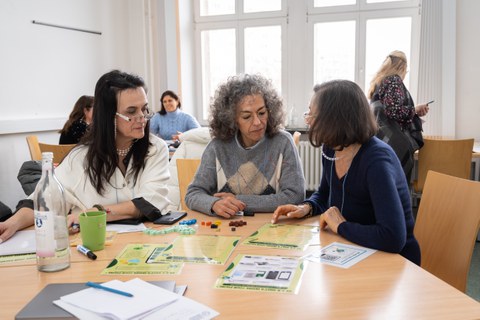 Drei Frauen sitzen an einem Tisch und diskutieren. Es liegen verschiedene Arbeitsmaterialien auf dem Tisch.