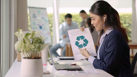 Eine Frau mit mit Kopfhörern im Ohr sitzt vor einem Laptop und hält ein Blatt mit dem Recycling-Symbol hoch.
