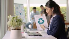 Eine Frau mit mit Kopfhörern im Ohr sitzt vor einem Laptop und hält ein Blatt mit dem Recycling-Symbol hoch.