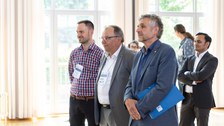 Das Foto zeigt drei Männer, die nebeneinander stehen und bei einer Vorstellungsrunde zuhören.