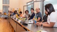 Das Foto zeigt mehrere Personen, die in einer Reihe an einem Konferenztisch sitzen mit Mikrofon an jedem Platz.