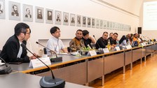 Das Foto zeigt mehrere Personen, die in einer Reihe an einem Konferenztisch sitzen mit Mikrofon an jedem Platz.