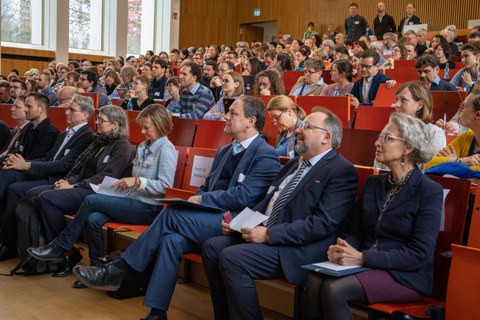 Foto: Personen in einem vollbesetzten Hörsaal blicken zur Bühne.