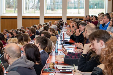Foto: Personen in einem vollbesetzten Hörsaal blicken zur Bühne.