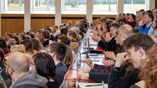 Foto: Personen in einem vollbesetzten Hörsaal blicken zur Bühne.