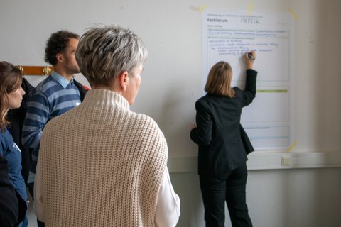 Foto: Mehrere Personen stehen vor einem Plakat, das an einer Wand befestigt ist. Eine Person schreibt etwas auf das Plakat.