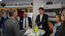 Foto: Mehrere Personen stehen um einen Tisch und unterhalten sich.