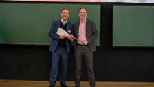 Foto: Zwei Personen stehen vor einer Tafel und halten ein Ruder in den Händen.