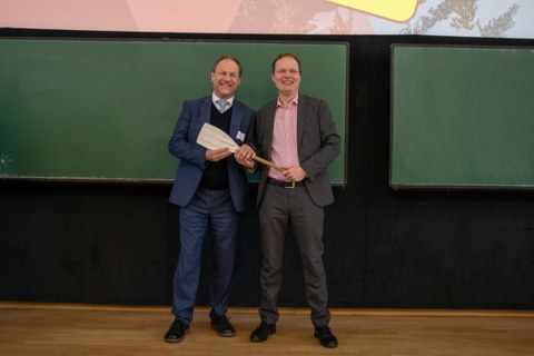 Foto: Zwei Personen stehen vor einer Tafel und halten ein Ruder in den Händen.