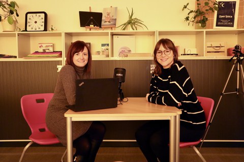 Auf dem Foto sind 2 Frauen zu sehen, die sich an einem Tisch im Lehr-Lern-Raum Inklusion gegenübersitzen. Auf dem Tisch steht ein Laptop und ein Podcastmikrofon. Die beiden Frauen lächeln in die Kamera.
