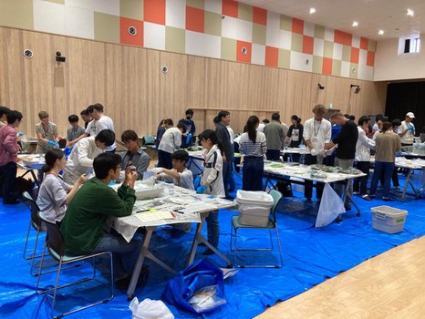 Gruppe von ca. 50 Studierenden, die in einem Saal an mehren großen Tischen zusammen arbeiten. 