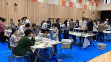 Gruppe von ca. 50 Studierenden, die in einem Saal an mehren großen Tischen zusammen arbeiten. 