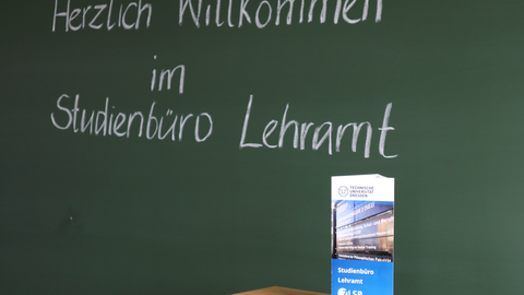 Das Foto zeigt eine Tafel, auf der "Herzlich Willkommen im Studienbüro Lehramt" geschrieben steht. Auf der Unterkante der Tafel steht eine Informationsbroschüre des Studienbüros Lehramt.