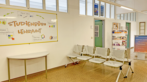 Bild des Ganges im 2. Obergeschoss im Seminargebäude 2. Auf einer großen Tafel steht "Studienbüro Lehramt". 2 Sitzbänke, Aufsteller mit Informationsflyern und eine offene Tür zum Front Desk sind zu sehen.