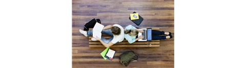 Das Foto zeigt zwei Studierende aus der Vogelperspektive. Sie sitzen Rücken an Rücken auf einer Bank und arbeiten. Um die Bank herum liegen Taschen und weitere Arbeitsmaterialien verteilt.
