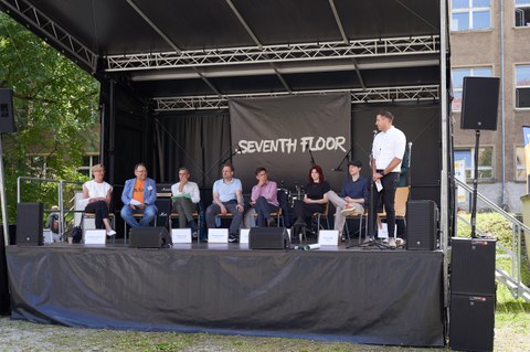 Diskussionsrunde auf einer Bühne mit 7 Diskutant:innen und einem Moderator