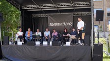 Diskussionsrunde auf einer Bühne mit 7 Diskutant:innen und einem Moderator