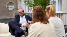 Ein Mann spricht mit zwei jungen Frauen auf einem Sofa. Der Mann sitzt den beiden Frauen dabei gegenüber.