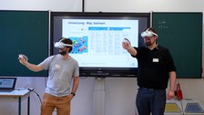 Zwei Männer die vor einer Schultafel stehen jeweils eine VR-Brille ausprobieren.