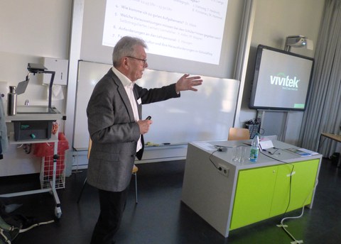 Bild, auf dem Prof. Dr. Körndle vor einer Tafel steht und einen Vortrag hält. Über der Tafel wird eine Präsentation an die Wand projiziert. Rechts im Bild sieht man eine aktive digitale Tafel.