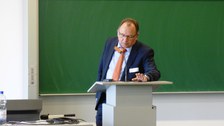 Bild, auf dem Prof. Dr. Gehrmann, Projektleiter des TUD-Sylber, vor einer Tafel an einem Pult mit Mikro steht.